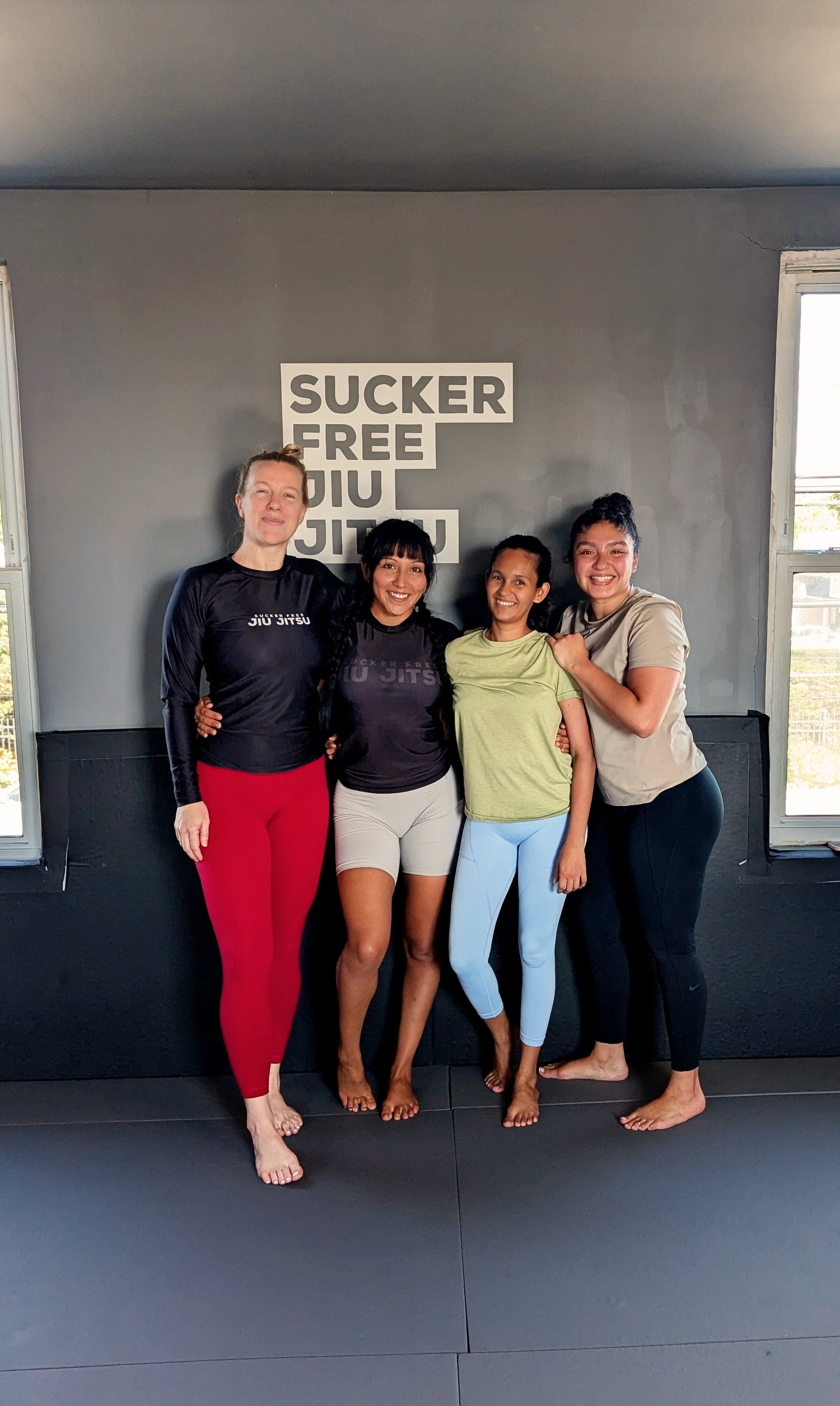 Coach Alli standing with three women students at the studio. They are standing in front of the Sucker Free Jiu Jitsu logo on the white and grey wall