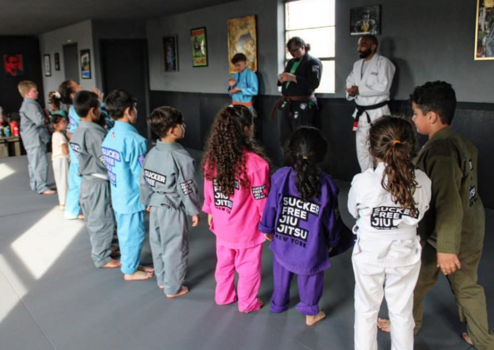 A group of children wearing colorful Jiu Jitsu gi stand in a line facing Coach Weiss, Coach Hynes and Coach Matty in the studio. The children's uniforms are in various colors including gray, light blue, pink, purple, white, and olive green, each printed with 'SUCKER FREE JIU JITSU' block logo on the back.